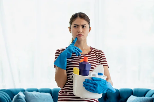 Stressed young Asian housewife wearing gloves and holding basket with cleaners and brushes feeling tired and frustrated, cleaning and washing concept