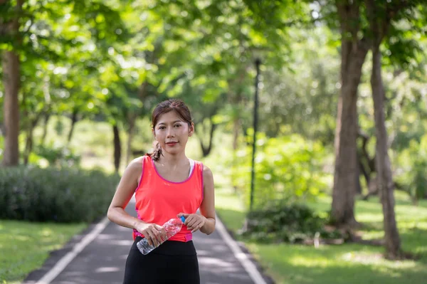 Happy Asian female runner standing outdoors holding water bottle in the park