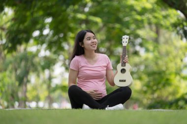 Asyalı sağlıklı hamile kadın parkta Ukulele oynayarak, anne sağlığı ve müzik yaparak eğleniyor ve dinleniyor.