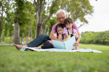 Mutlu Asyalı aile çocukları büyükbabasıyla parkta kitap okuyor.