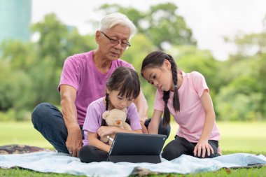 Mutlu Asyalı aile çocukları büyükbabasıyla parkta tablet kullanıyor.