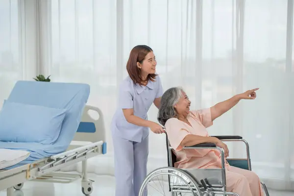 Asian nurse pushing senior female patient in a wheelchair at the hospital ward, elderly care concept