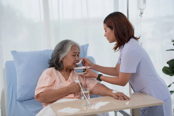 Young Asian nurse feeding breakfast to senior woman patient on bed in hospital ward, elderly care concept