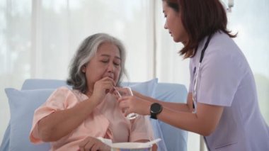 Asyalı hemşire hastane koğuşunda yatağında içmesi için yaşlı bir kadına bir bardak su veriyor.