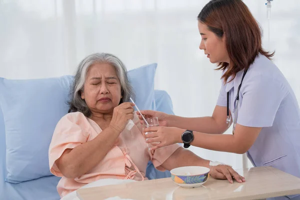 Asian nurse giving glass of water to elderly woman patient to drink on bed at hospital ward