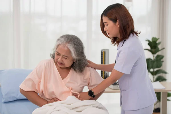 Asian nurse helping a senior Asian female patient with stomach ache on a bed in hospital ward with diagnosis or treatment, treating elderly woman