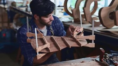 Luthier bir gitar yaratıyor ve geleneksel olarak aletleri kullanıyor. Yapım aşamasında olan bir gitarın gövdesine kelepçe takıyor. Yapıştırıcı yapışkanlığını geliştiriyor.