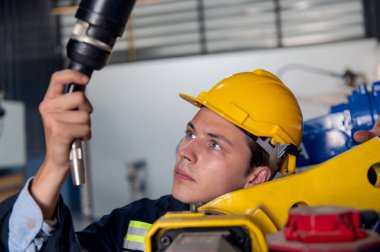 Kask güvenlik teftişi yapan bir mühendis endüstriyel fabrikada uzaktan kumandalı bir robot kolu kaynak makinesini kontrol ediyor. Teknisyen tamirat işçisi elektronik işlemleri kontrol ediyor.