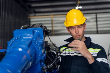 Kask güvenlik teftişi yapan bir mühendis endüstriyel fabrikada uzaktan kumandalı bir robot kolu kaynak makinesini kontrol ediyor. Teknisyen tamirat işçisi elektronik işlemleri kontrol ediyor.
