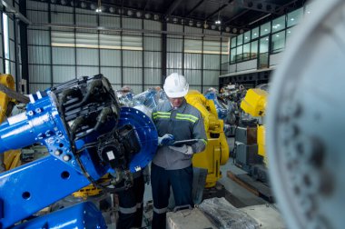 Kask güvenlik teftişi yapan bir mühendis endüstriyel fabrikada uzaktan kumandalı bir robot kolu kaynak makinesini kontrol ediyor. Teknisyen tamirat işçisi elektronik işlemleri kontrol ediyor.