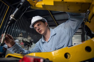 Kask güvenlik teftişi yapan bir mühendis bir endüstriyel fabrikada uzaktan kumandalı bir robot kol kaynak makinesini kontrol ediyor.