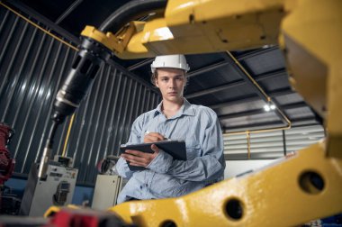 Robotik kol ve işletim makinesinin yanında duran miğfer güvenlikli mühendis. Teknisyen, tamir onarım elektronik işlemleri için kontrol ediyor.