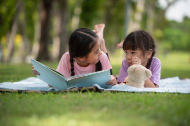 Mutlu Asyalı aile çocukları, abla ve kız kardeş parkta birlikte kitap okuyorlar.