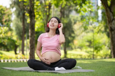 Young Asian pregnant woman enjoys listening to music with headphones in the park, happy and relaxing time