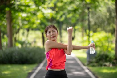 Mutlu Asyalı kadın koşucu dışarıda ısınıyor. Parkta su şişesi tutuyor.