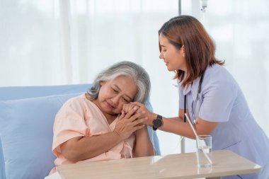 Genç Asyalı kadın hemşire, yaşlı bayana yardım ediyor ve hastanedeki bir yatakta hastayı cesaretlendiriyor.