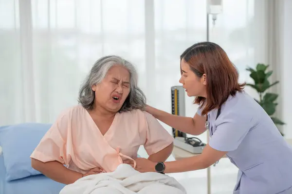 stock image Asian nurse helping a senior Asian female patient with stomach ache on a bed in hospital ward with diagnosis or treatment, treating elderly woman