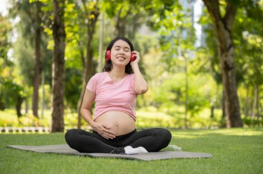 Young Asian pregnant woman enjoys listening to music with headphones in the park, happy and relaxing time