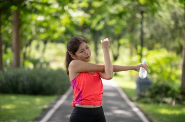 Mutlu Asyalı kadın koşucu dışarıda ısınıyor. Parkta su şişesi tutuyor.