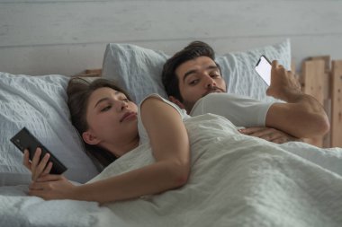Yatakta cep telefonu kullanan genç bir çift sırt sırta oturarak, ilişki ve iletişim sorunları, internet sosyal ağ bağımlılığı