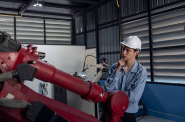 Robotik kol ve işletim makinesinin yanında duran miğfer güvenlikli mühendis. Teknisyen, tamir onarım elektronik işlemleri için kontrol ediyor.