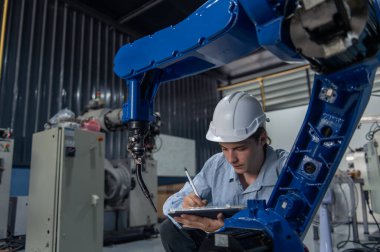 Robotik kol ve işletim makinesinin yanında duran miğfer güvenlikli mühendis. Teknisyen, tamir onarım elektronik işlemleri için kontrol ediyor.