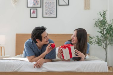 Genç Asyalı çift yatakta yatıp kırmızı kalp ve hediye kutusuyla birlikte vakit geçiriyorlar Sevgililer Günü 'nü kutluyorlar, mutlu aile kavramları.