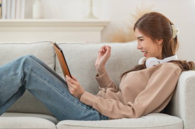 Genç Asyalı kadın, evdeki modern oturma odasında dijital tablet kullanırken kulaklıkla müzik dinlemeyi seviyor, mutlu ve rahatlatıcı bir zaman.