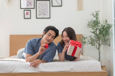 Genç Asyalı çift yatakta yatıp kırmızı kalp ve hediye kutusuyla birlikte vakit geçiriyorlar Sevgililer Günü 'nü kutluyorlar, mutlu aile kavramları.