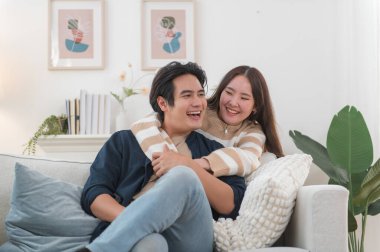 Mutlu genç Asyalı çift sarılıyor, rahat bir kanepede birlikte oturuyor, aşırı mutlu bir şekilde gülen kadın ve erkek eğleniyor, boş vakitlerin tadını çıkarıyor, evdeki oturma odasında kanepede dinleniyor, iyi bir ilişki içinde.