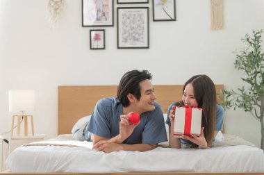 Genç Asyalı çift yatakta yatıp kırmızı kalp ve hediye kutusuyla birlikte vakit geçiriyorlar Sevgililer Günü 'nü kutluyorlar, mutlu aile kavramları.