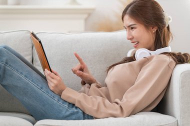 Genç Asyalı kadın, evdeki modern oturma odasında dijital tablet kullanırken kulaklıkla müzik dinlemeyi seviyor, mutlu ve rahatlatıcı bir zaman.