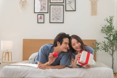 Genç Asyalı çift yatakta yatıp kırmızı kalp ve hediye kutusuyla birlikte vakit geçiriyorlar Sevgililer Günü 'nü kutluyorlar, mutlu aile kavramları.