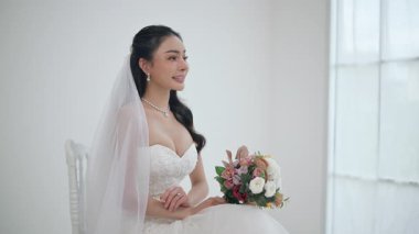 Düğün, gelinlik içinde güzel bir kanepede oturuyor beyaz stüdyo içinde evdeki gibi, genç çekici Asyalı kadın ışıl ışıl gülümse