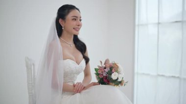 Düğün, gelinlik içinde güzel bir kanepede oturuyor beyaz stüdyo içinde evdeki gibi, genç çekici Asyalı kadın ışıl ışıl gülümse