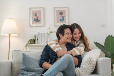 Mutlu genç Asyalı çift sarılıyor, rahat bir kanepede birlikte oturuyor, aşırı mutlu bir şekilde gülen kadın ve erkek eğleniyor, boş vakitlerin tadını çıkarıyor, evdeki oturma odasında kanepede dinleniyor, iyi bir ilişki içinde.