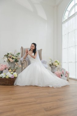 Düğün, güzel elbiseli gelin, beyaz stüdyo içinde kapalı koltukta oturuyor, genç çekici Asyalı kadın ışıl ışıl gülümse.