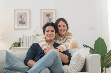 Mutlu genç Asyalı çift sarılıyor, rahat bir kanepede birlikte oturuyor, aşırı mutlu bir şekilde gülen kadın ve erkek eğleniyor, boş vakitlerin tadını çıkarıyor, evdeki oturma odasında kanepede dinleniyor, iyi bir ilişki içinde.