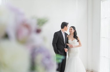 Düğün kıyafetleri içinde mutlu Asyalı gelin ve damat gülümseyip birbirlerine bakıyorlar, evin içinde parlak pencere ışığında duruyorlar ve ön planda çiçek süslemeleri var. Düğün fotoğrafçılığı için romantik çift portresi.