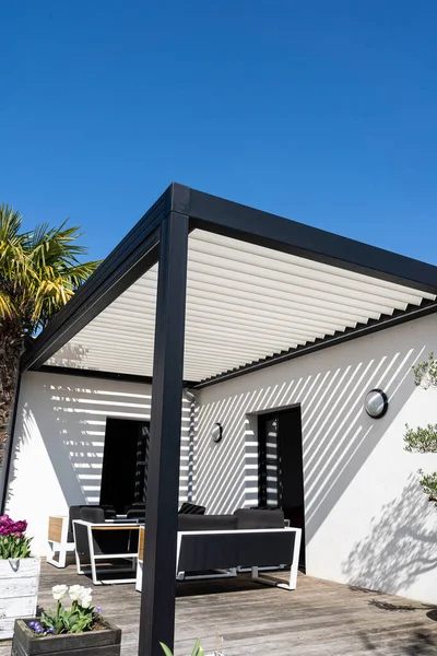 Çevre dostu biyo-mekanik alüminyum pergola gölge yapısı, tente ve teras çatısı, bahçe salonu, sandalyeler, bahçe düzeniyle çevrili metal ızgara.