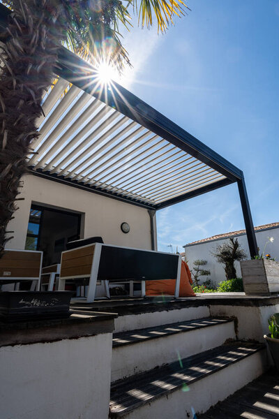 realistic photo modern design bioclimatic pergola with aluminum slats which stands near a modern house with swimming pool , with dining area , rich in vegetation