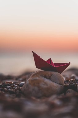 Renkli bir origami teknesi çakıl taşı plajında dinleniyor. Gün batımında okyanus dalgaları yavaşça sahili öpüyor.