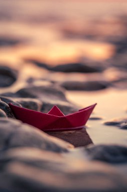 Renkli bir origami teknesi çakıl taşlı bir plajda seyrediyor. Gün batımında okyanus dalgaları yavaşça sahili öpüyor.