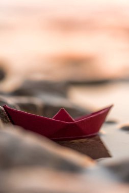 Renkli bir origami teknesi çakıl taşlı bir plajda seyrediyor. Gün batımında okyanus dalgaları yavaşça sahili öpüyor.