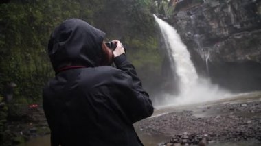 Şelalenin önündeki kadın fotoğrafçı yağmurun altında ve ormanın ortasında. Bali, Endonezya. Yüksek kaliteli FullHD yavaş çekim görüntüleri.