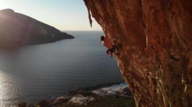 Günbatımında Grande Grotta mağarasının sert ve dik bir bölümüne tırmanan dağcı ve Yunanistan 'ın Kalymnos adasının arkasındaki Telendos adası ve kıyı şeridi. Yüksek kalite 4k görüntü.