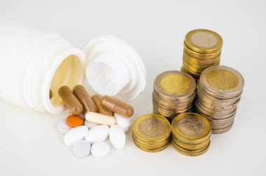 Medicines and tablets, coming out of a container, with stacks of coins next to them. Cost of medicines and price increase. 