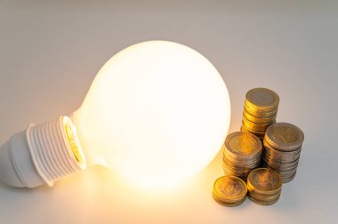 Light bulb turned on, with stacks of coins next to it. Rising electricity tariffs, energy dependency, energy sources and energy supplies.
