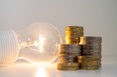 Light bulb turned on, with stacks of coins next to it. Rising electricity tariffs, energy dependency, energy sources and energy supplies.