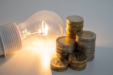 Light bulb turned on, with stacks of coins next to it. Rising electricity tariffs, energy dependency, energy sources and energy supplies.